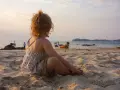 Liten flicka sitter på stranden och tittar ut över havet. Det är varmt på vintern, med båtar i bakgrunden och en vacker solnedgång.