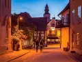 Två personer promenerar på en kullerstensgata i Båstad under Stockholmsveckan. Kvällen är ljum, och husen längs gatan lyser varmt.