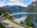 En bil kör längs en slingrande väg vid en fjord, med snöklädda berg i bakgrunden. En perfekt bild för en semester i Norge.