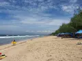 En man med en gul surfbräda går längs Kuta Beach. Vågor rullar in mot stranden där folk solar och surfar.