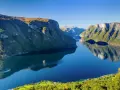 En båt färdas på en spegelblank fjord i Norge, omgiven av gröna bergssluttningar. En tågresa genom detta landskap vore en dröm.