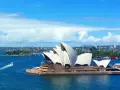 Sydney stad med operahuset i förgrunden och en båt på det blå vattnet.