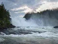 Vackra vattenfall dånar nerför klipporna, omgivna av en tät grön skog under en dramatisk molnhimmel.