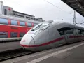 Ett modernt ICE-tåg står redo vid perrongen, perfekt för en resa med tåg till Tyskland. Bredvid syns ett rött dubbeldäckartåg.
