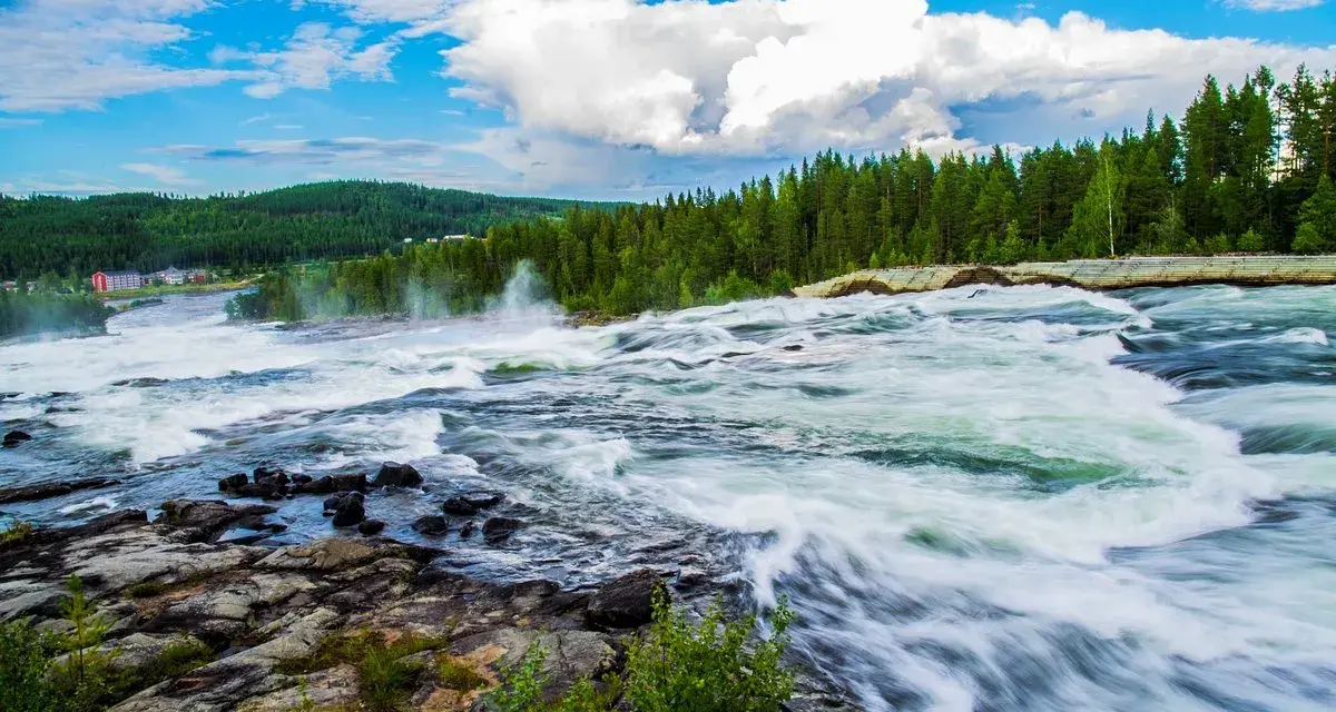 Vattenfall i Luleå med forsande vatten, grönskande skog och en blå himmel med vita moln.