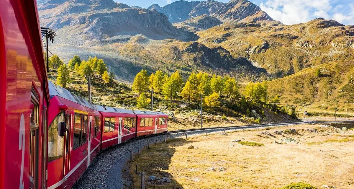 Ett rött tåg slingrar sig genom ett höstfärgat landskap med berg i bakgrunden. Perfekt för att tågluffa i Sverige och uppleva naturens skönhet.