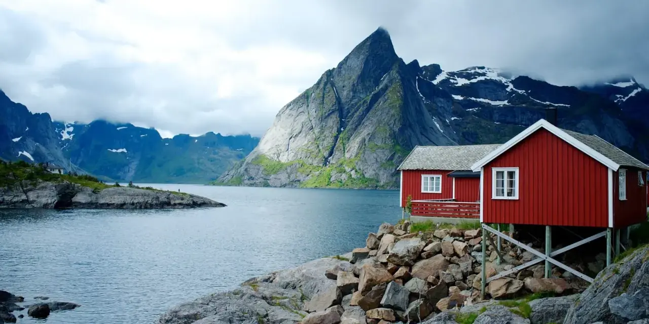 Två röda stugor vid en sjö i Norge, med dramatiska berg i bakgrunden. Perfekt för den som vill vandra i Norge.