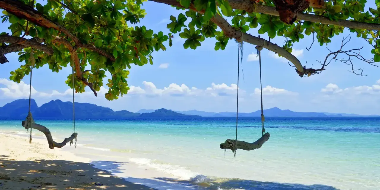 Gungor hänger från ett träd vid en strand i Thailand. Vackert turkost vatten och berg i bakgrunden. En idyllisk plats, trots att det är regnperiod.