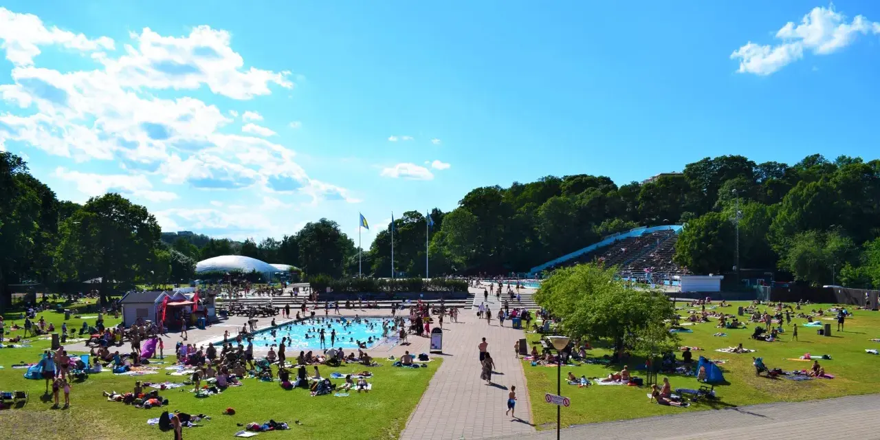En solig dag vid en populär badplats i Stockholm. Människor solar, badar och njuter av sommaren på gräsmattor och i poolen.