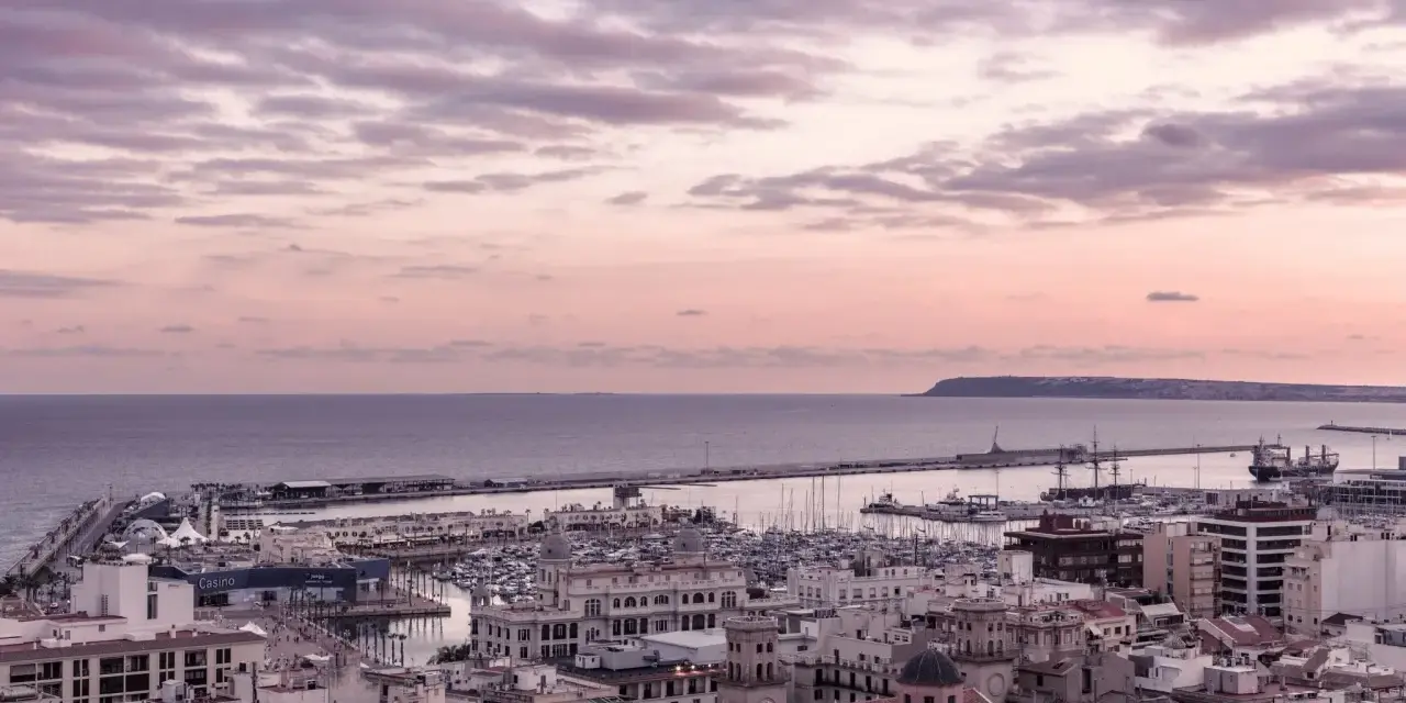 En vacker solnedgång över hamnen i Alicante i december. Molnen färgas i rosa och lila över havet och staden.