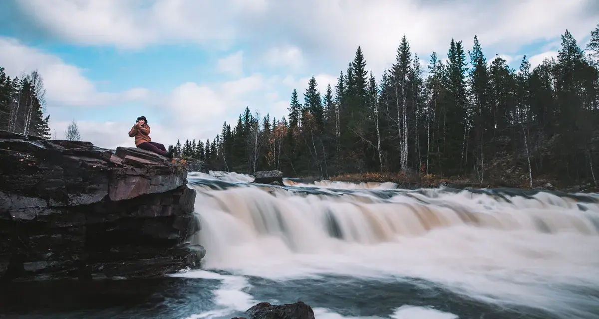 Person sitter på en klippa vid ett forsande vattenfall i Dalarna, ett perfekt utflyktsmål.