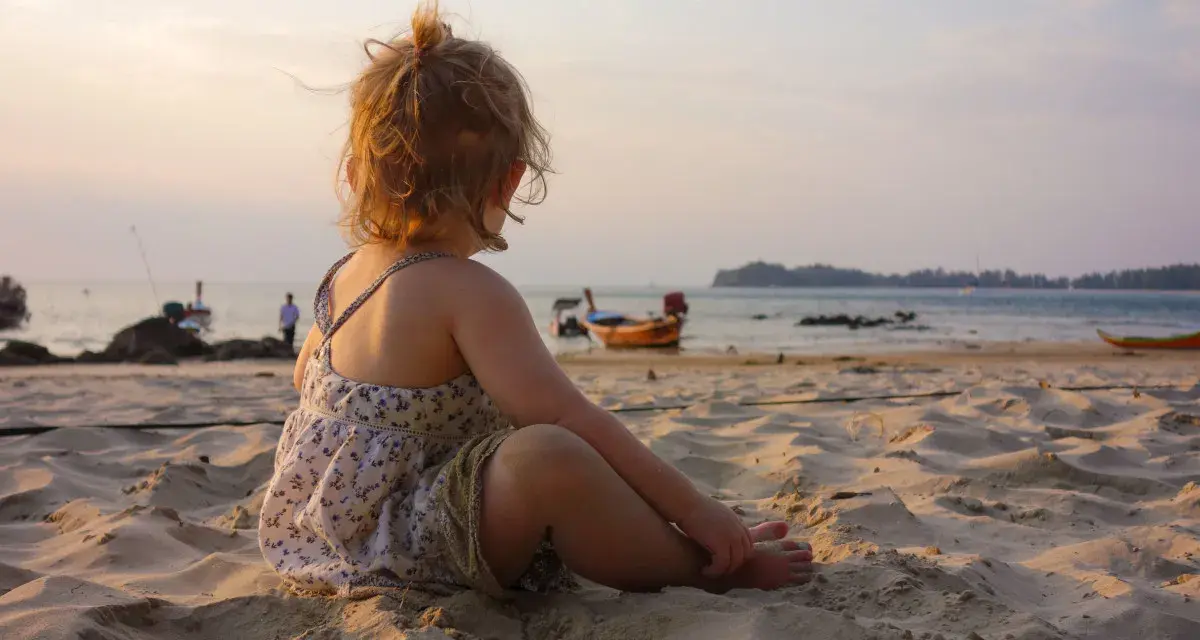 Liten flicka sitter på stranden och tittar ut över havet. Det är varmt på vintern, med båtar i bakgrunden och en vacker solnedgång.