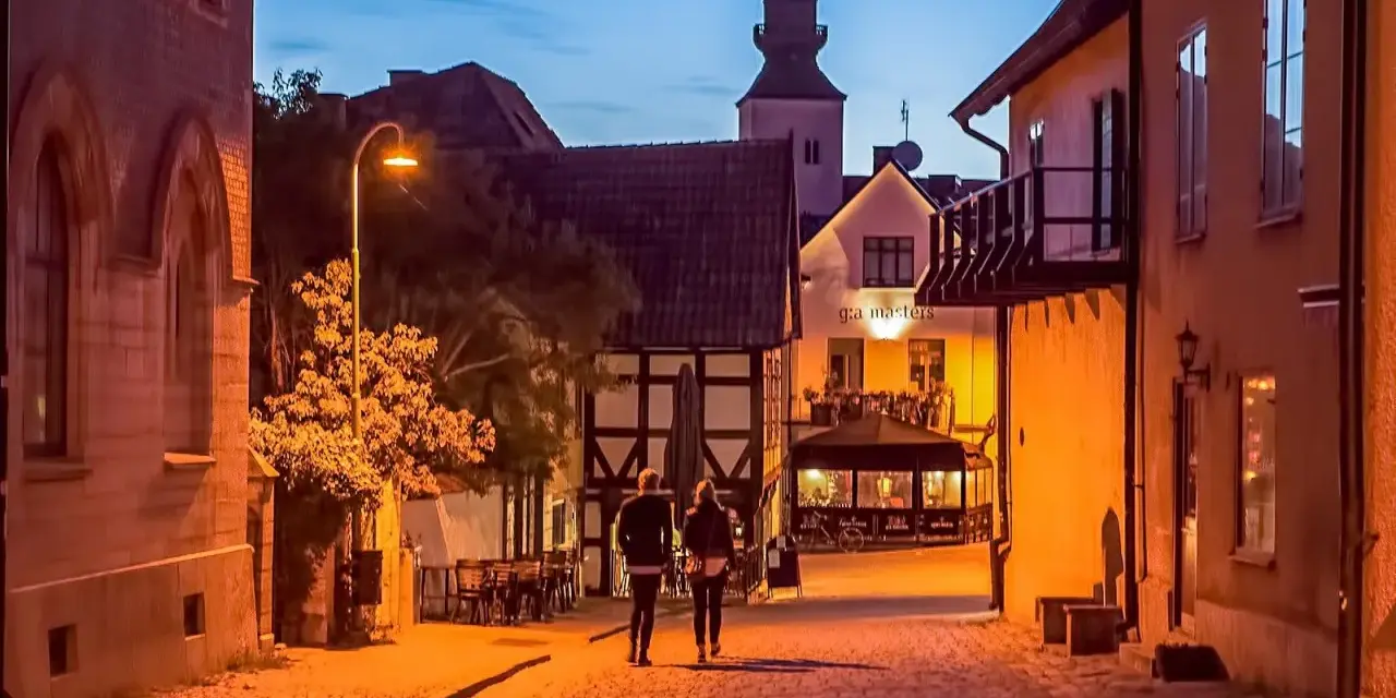 Två personer promenerar på en kullerstensgata i Båstad under Stockholmsveckan. Kvällen är ljum, och husen längs gatan lyser varmt.