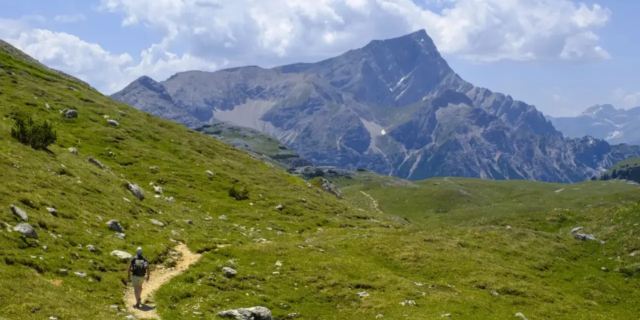 Vandrare på en stig i Dolomiterna, omgiven av gröna sluttningar och majestätiska berg under en molnig himmel.