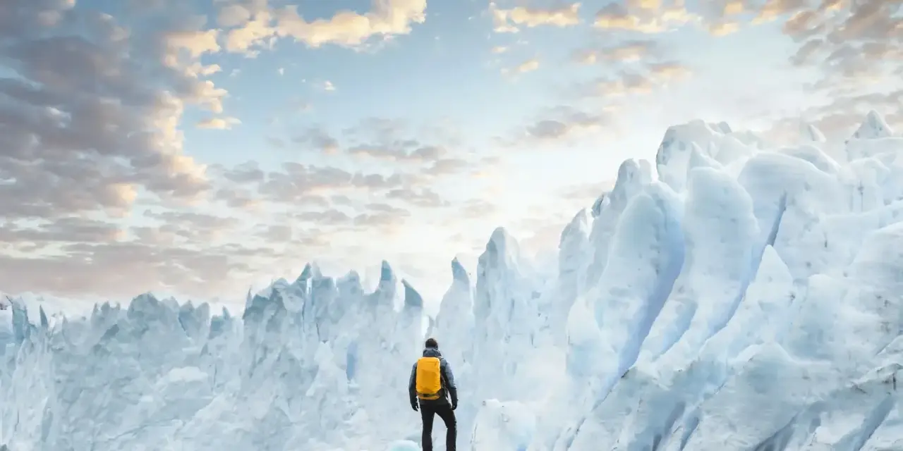 En person med gul ryggsäck står på stenar framför en enorm glaciär. Ett perfekt resmål i januari för den äventyrlige.