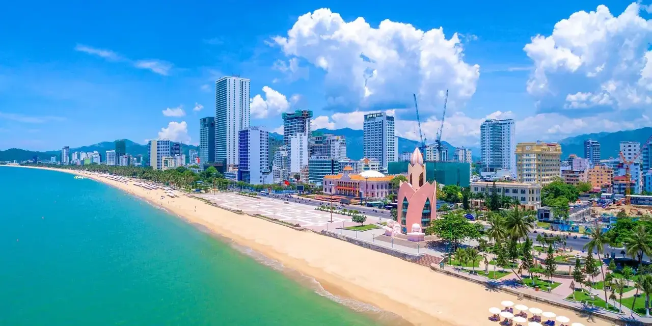 En vidsträckt strand med turkost vatten och en stadssilhuett i bakgrunden. Nha Trang beach är en populär destination.