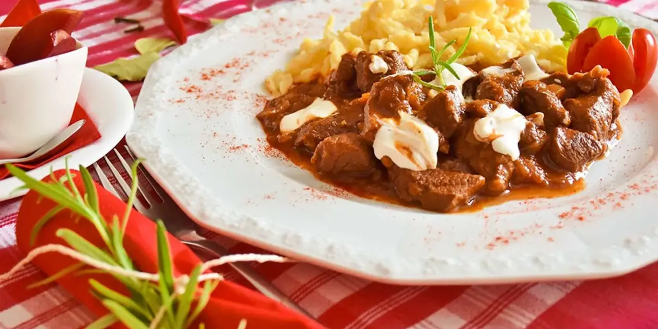 En smak av mat i Budapest: mustig gulasch med nokedli, garnerad med gräddfil och en rosmarinkvist. En röd chili och en tomatblomma fulländar upplägget.
