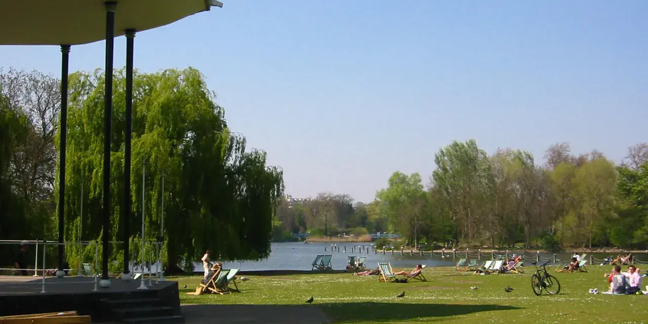 En solig dag i en park i London. Människor vilar på solstolar vid en sjö, med en grönskande trädgård och en paviljong i förgrunden.
