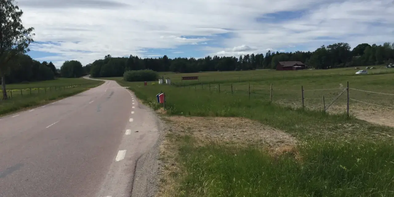 Vacker väg kantad av grönska, perfekt för att utforska Sveriges cykelleder. En röd lada skymtar i bakgrunden.