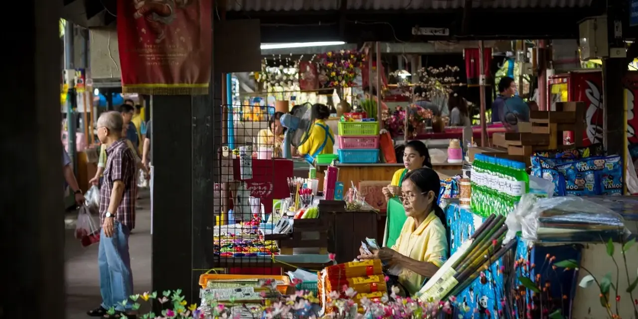 En livlig marknad i Bangkok med försäljare och kunder bland färgglada varor och blommor.