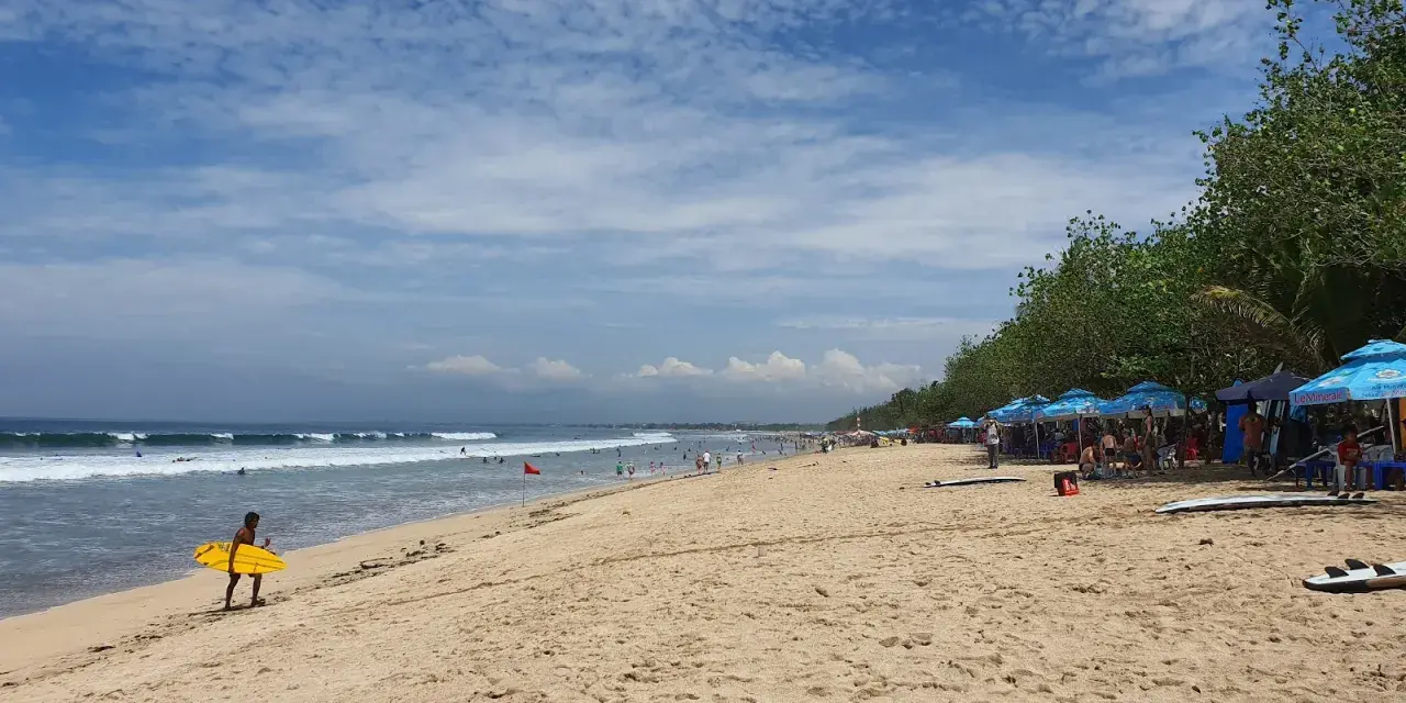 En man med en gul surfbräda går längs Kuta Beach. Vågor rullar in mot stranden där folk solar och surfar.