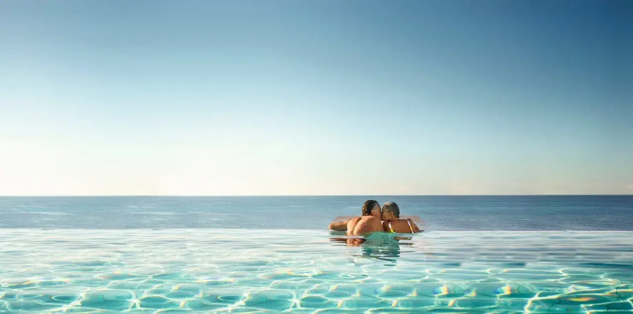 Ett par kysser varandra i en infinitypool med havet i bakgrunden. Perfekt för romantiska resmål.