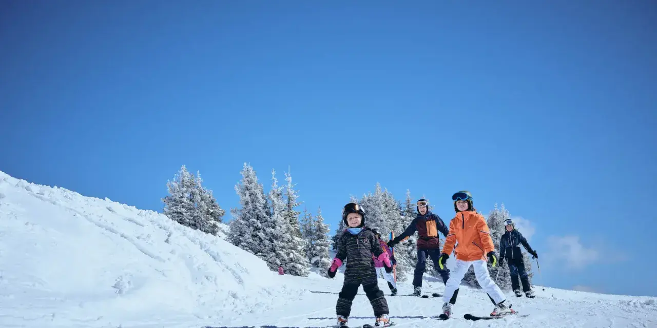 Familj njuter av skidåkning i mars. Barn och vuxna i skidkläder på en snöig backe med träd i bakgrunden. Ett perfekt resmål mars för vinteraktiviteter.