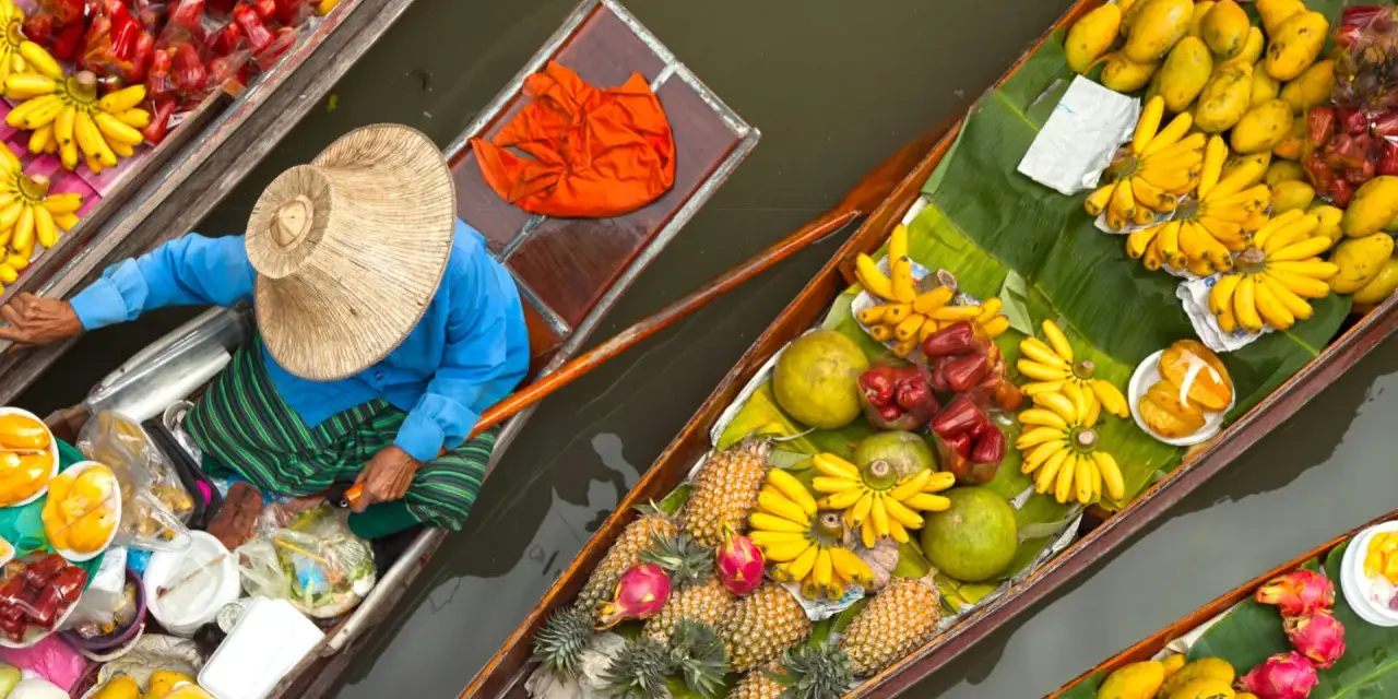En säljare i en båt på en flytande marknad i Thailand, omgiven av ett överflöd av färsk frukt som bananer, mango och ananas.