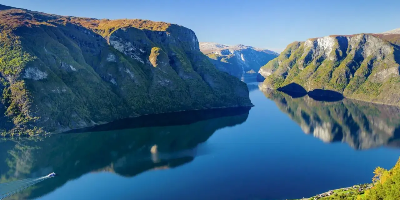 En båt färdas på en spegelblank fjord i Norge, omgiven av gröna bergssluttningar. En tågresa genom detta landskap vore en dröm.