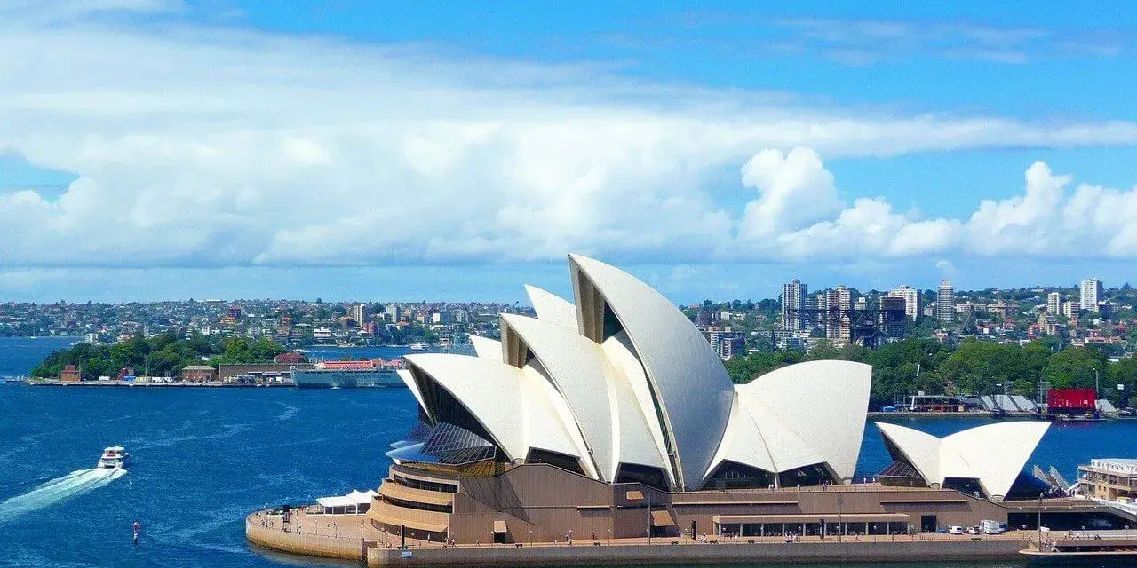 Sydney stad med operahuset i förgrunden och en båt på det blå vattnet.