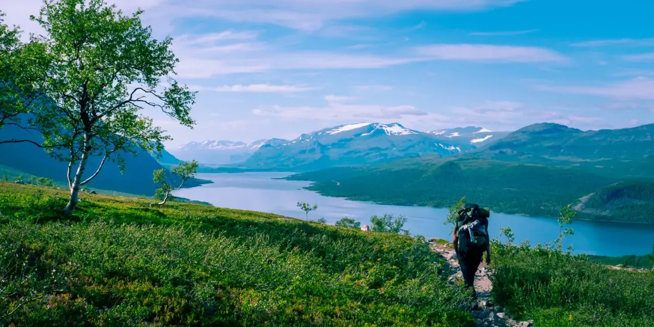 Vandrare med stor ryggsäck på en stenstig, njuter av storslagna vyer över sjö och snöklädda berg. Perfekt för vandringsleder Norrland.