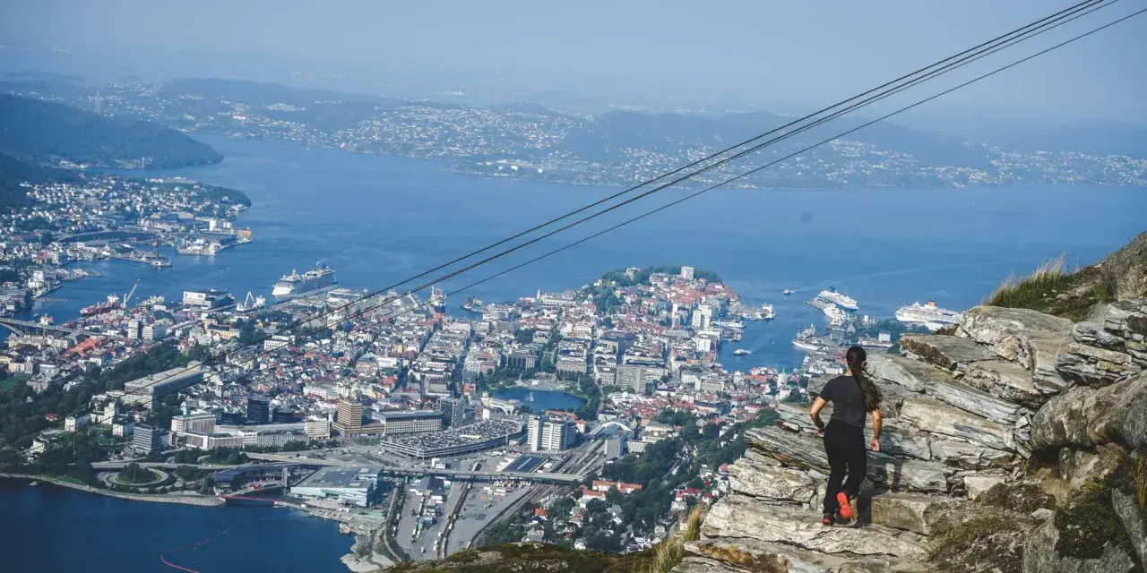 En kvinna står på en klippa och blickar ut över en stad vid havet, med berg i bakgrunden.