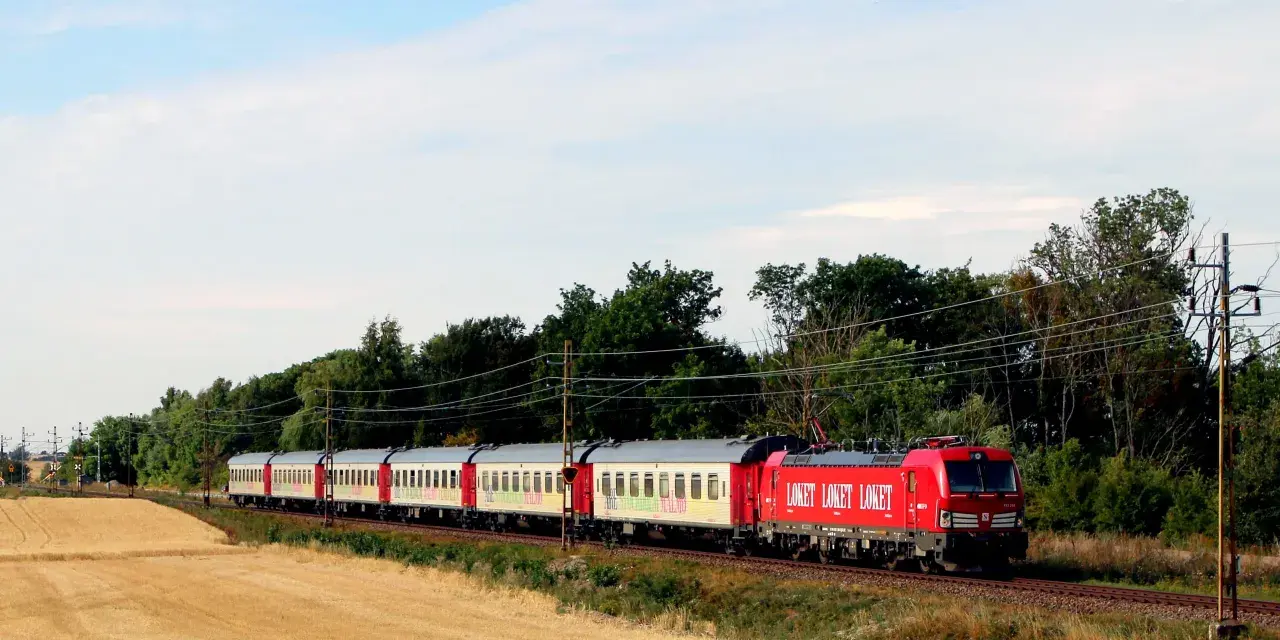 Ett rött lok drar ett långt tåg med vita vagnar genom ett landskap med guldigt höstfält och gröna träd. Kanske är det ett nattåg till Berlin.