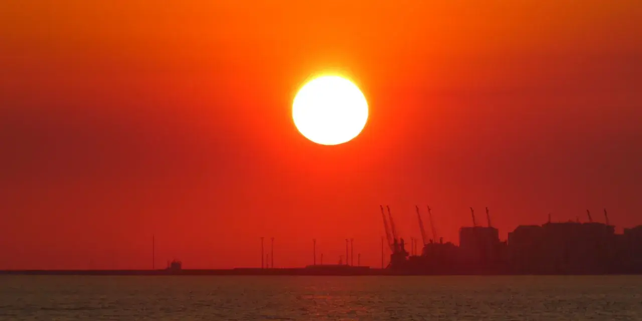 En glödande sol går ner över havet, med silhuetter av hamnkranar och byggnader i fjärran. En av de fina solnedgångar som får en att drömma sig bort.