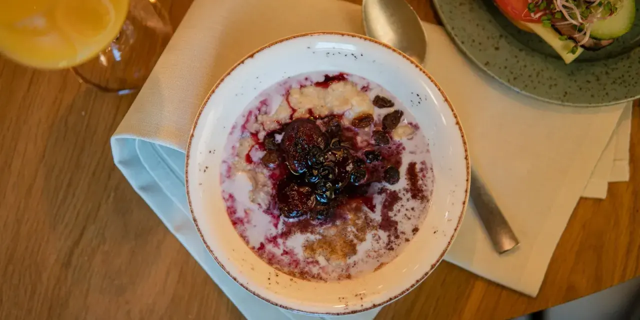 En skål med gröt toppad med bär och russin, en del av en kroatisk frukost. Bredvid syns en smörgås och ett glas juice.