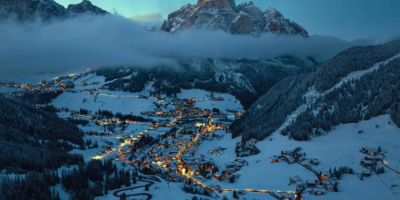 En snötäckt by i Italien på vintern, upplyst av tusentals ljus. Majestätiska berg reser sig bakom, inbäddade i dimma.