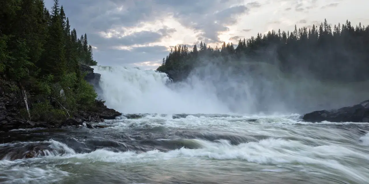 Vackra vattenfall dånar nerför klipporna, omgivna av en tät grön skog under en dramatisk molnhimmel.