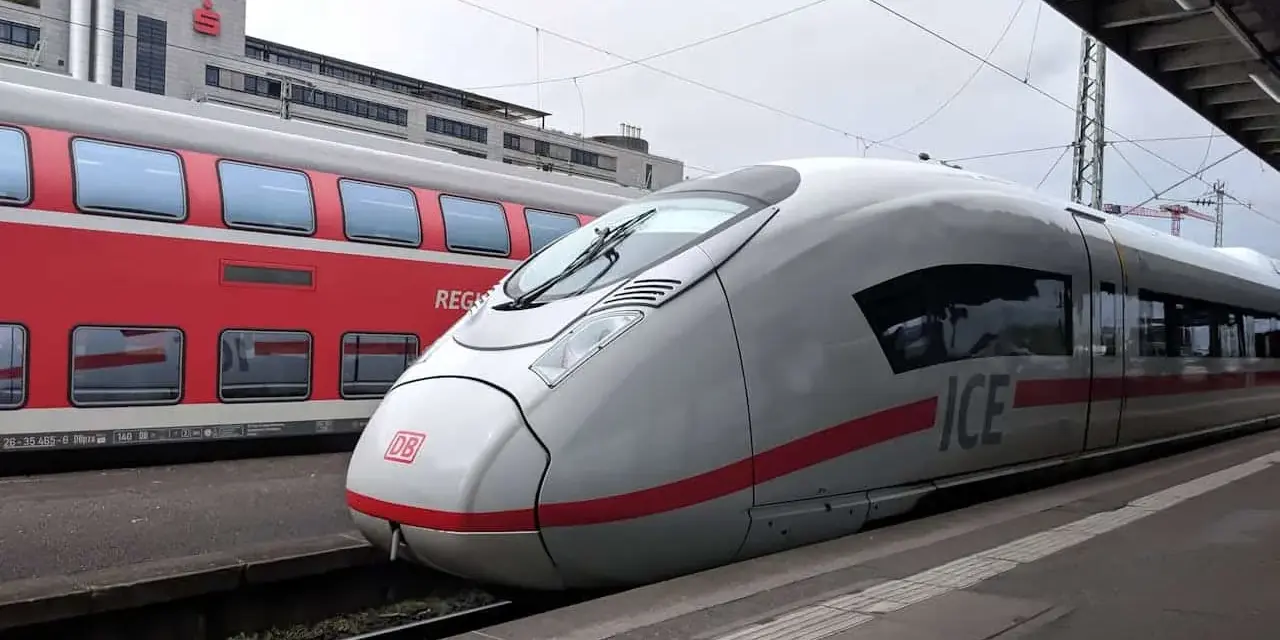 Ett modernt ICE-tåg står redo vid perrongen, perfekt för en resa med tåg till Tyskland. Bredvid syns ett rött dubbeldäckartåg.