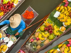 En säljare i en båt på en flytande marknad i Thailand, omgiven av ett överflöd av färsk frukt som bananer, mango och ananas.