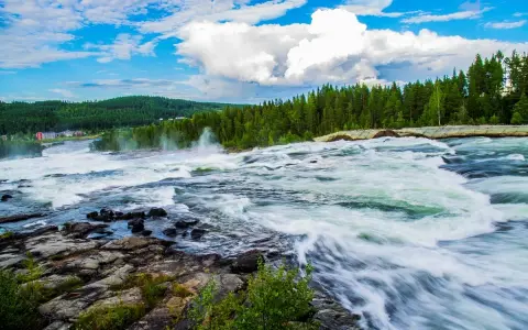 Vattenfall i Luleå med forsande vatten, grönskande skog och en blå himmel med vita moln.
