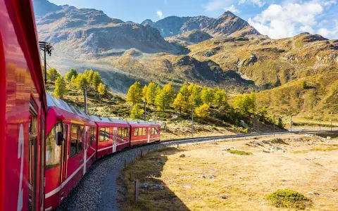 Ett rött tåg slingrar sig genom ett höstfärgat landskap med berg i bakgrunden. Perfekt för att tågluffa i Sverige och uppleva naturens skönhet.