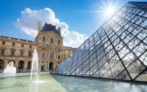 Louvren, ett berömt konstmuseum i Paris, med sin ikoniska glaspyramid och fontäner i förgrunden under en solig himmel.