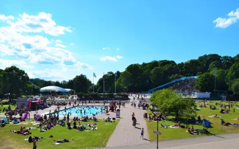 En solig dag vid en populär badplats i Stockholm. Människor solar, badar och njuter av sommaren på gräsmattor och i poolen.