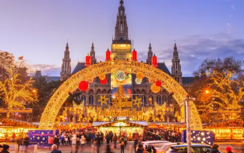 En magisk julmarknad i Wien, med Rådhuset i bakgrunden, gnistrar av tusentals ljus. En båge av gyllene ljusslingor välkomnar besökare till den livliga julmarknaden.