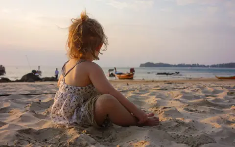 Liten flicka sitter på stranden och tittar ut över havet. Det är varmt på vintern, med båtar i bakgrunden och en vacker solnedgång.