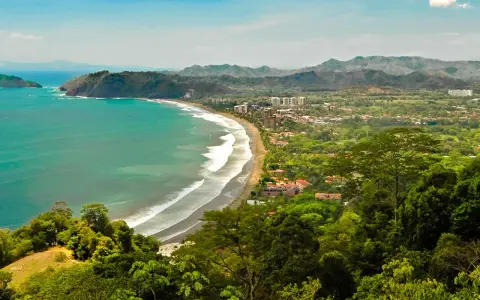 Vacker kustlinje med turkost hav, vågor som slår mot en sandstrand och frodig grönska som omger en stad. En dröm om en costa rica strand.