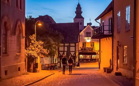 Två personer promenerar på en kullerstensgata i Båstad under Stockholmsveckan. Kvällen är ljum, och husen längs gatan lyser varmt.