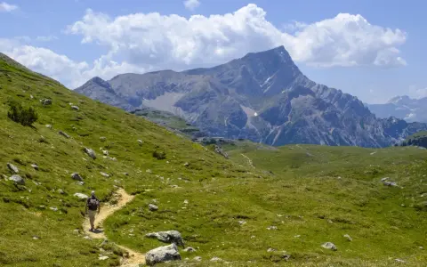 Vandrare på en stig i Dolomiterna, omgiven av gröna sluttningar och majestätiska berg under en molnig himmel.