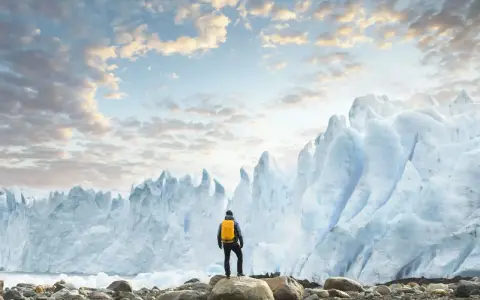 En person med gul ryggsäck står på stenar framför en enorm glaciär. Ett perfekt resmål i januari för den äventyrlige.