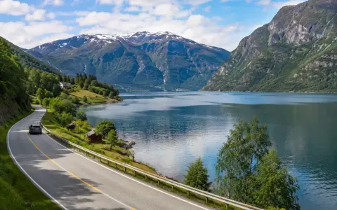 En bil kör längs en slingrande väg vid en fjord, med snöklädda berg i bakgrunden. En perfekt bild för en semester i Norge.